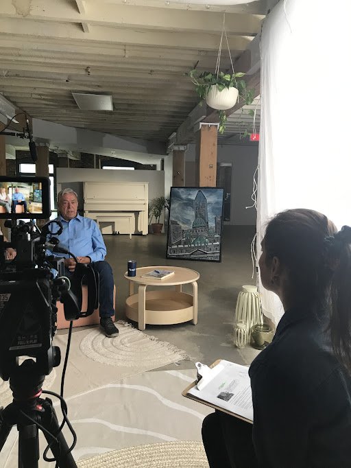 Un homme en chemise bleue assis dans un studio avec un caméraman filmant, une femme intervieweuse prenant des notes, et une peinture de gratte-ciel en arrière-plan.