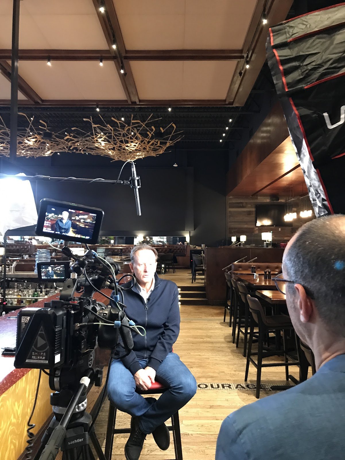 Un homme est en train d'être interviewé dans un restaurant ou un bar, avec un caméraman en face de lui, et un éclairage de tournage visible.