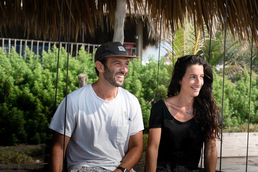 Un homme et une femme assis sur une balançoire sous un abri en chaume, souriant, avec des plantes vertes en arrière-plan.