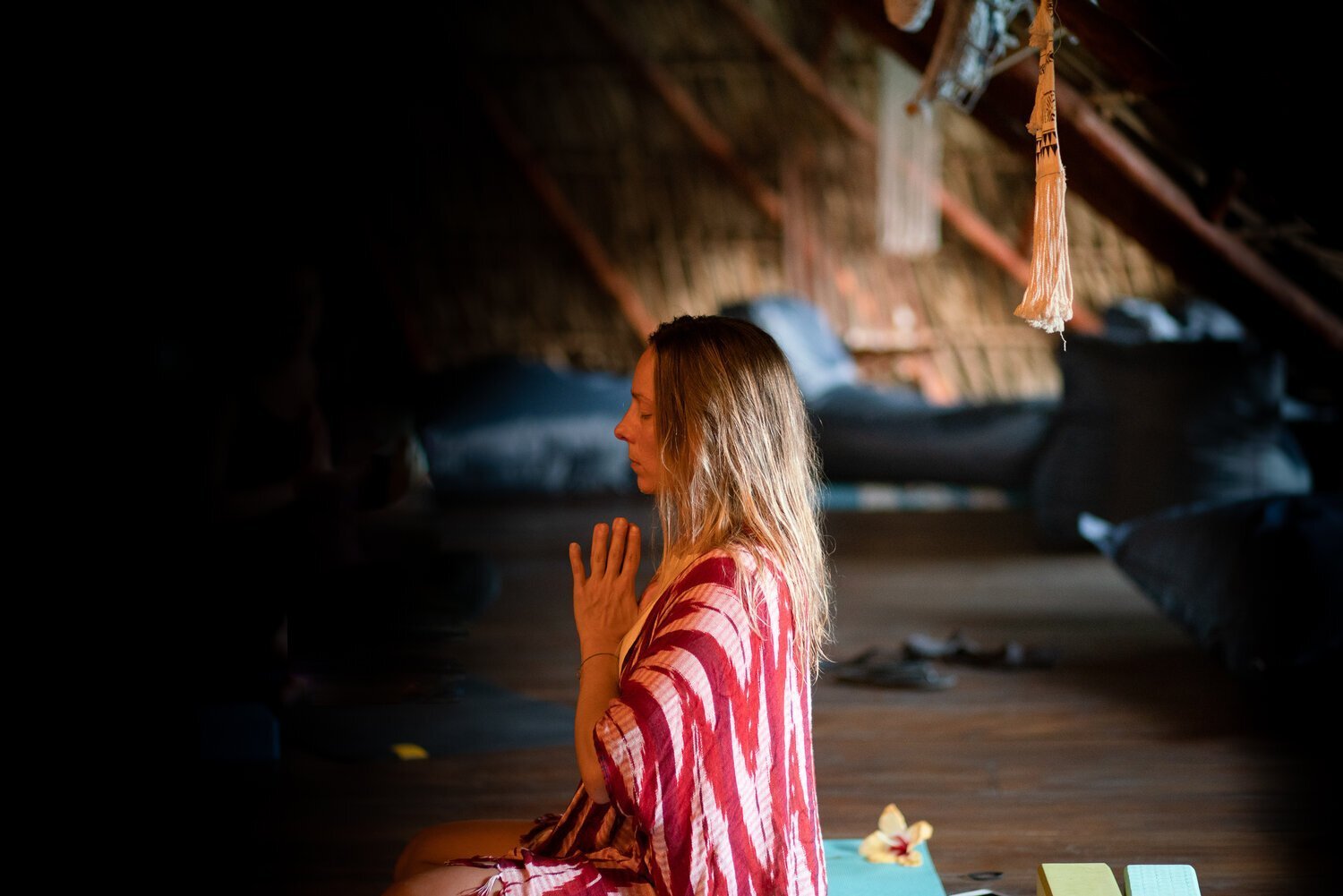 Femme méditant dans une salle sombre, portant une robe rayée rouge et blanche, avec les mains jointes près de son visage.