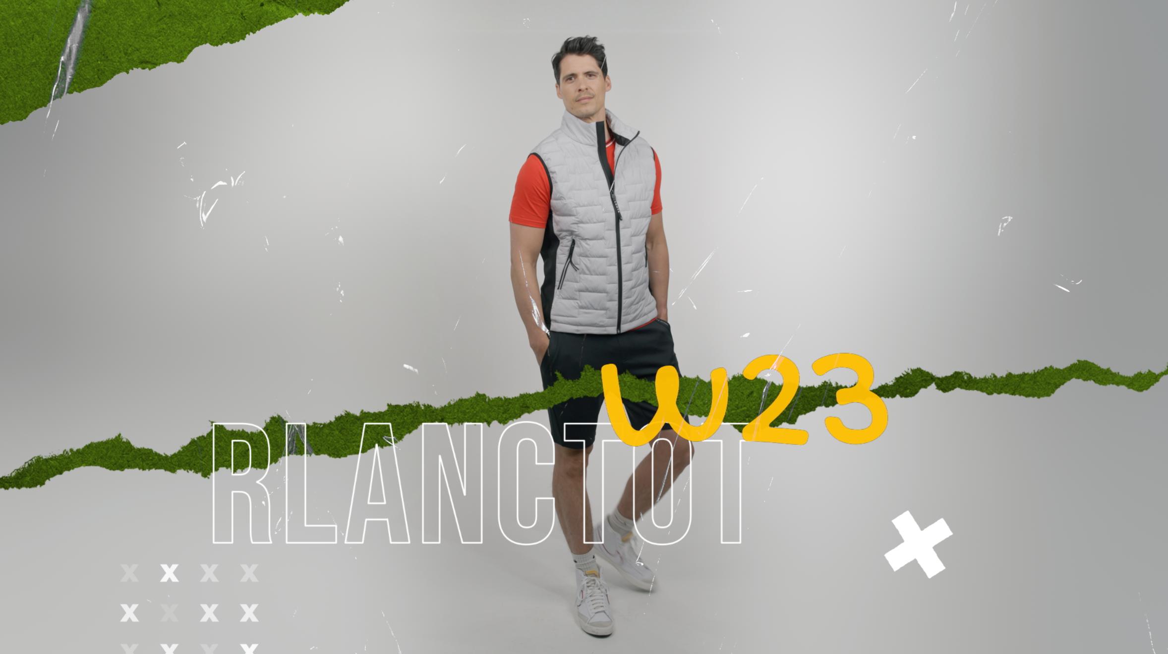 Homme portant une veste gris clair, t-shirt rouge, pantalon noir et chaussures de sport, posé devant un fond gris avec des éléments graphiques verts, blancs, jaunes et des textes "RLANCER W23" et des motifs en croix.