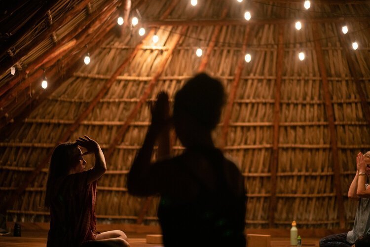 Des personnes pratiquant du yoga dans une pièce éclairée par des guirlandes lumineuses suspendues, avec un plafond en matériaux naturels.