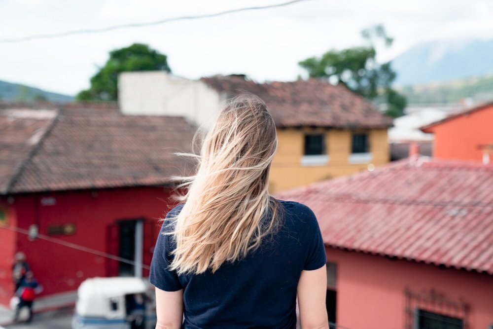 Une personne de dos avec de longs cheveux blonds, portant un t-shirt bleu, regardant des maisons aux toits en tuiles rouges dans un environnement urbain.